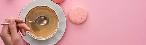 Vista cortada da mulher segurando colher de prata acima da xícara de café perto de deliciosos macaroons franceses no fundo rosa, tiro panorâmico — Fotografia de Stock