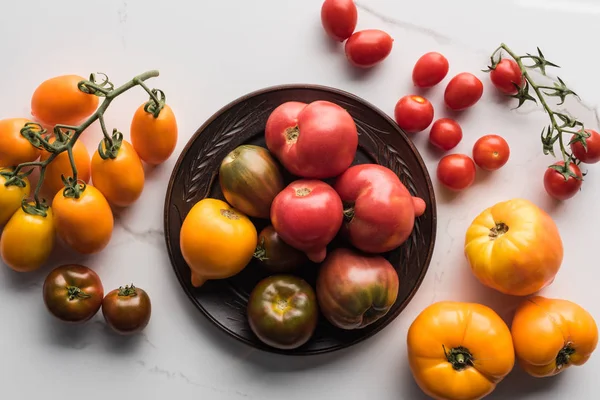 Vue du dessus des tomates sur assiette en bois près des tomates éparpillées sur la surface de marbre blanc — Photo de stock