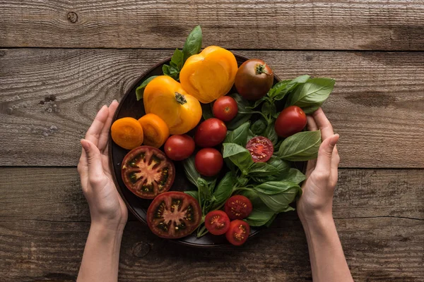 Cropped view of woman holding plate with spinach and sliced tomatoes on wooden table — Stock Photo