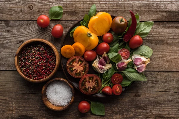 Vue du dessus de l'assiette avec épinards, ail et tomates tranchées près des bols avec poivre et sel sur la table en bois — Photo de stock