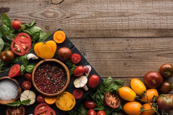 Vista dall'alto di vassoio nero con pepe e sale in ciotole, peperoncino, pomodori a fette e aglio vicino a verdure sparse sulla tavola di legno — Foto stock