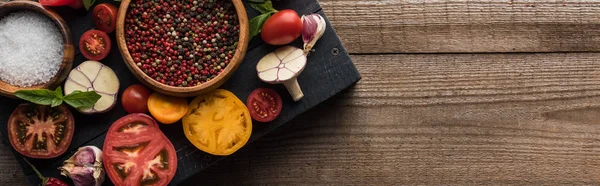 Panoramic shot of black tray with bowls with pepper and salt near chilli pepper, spinach, sliced tomatoes and garlic on wooden table — Stock Photo