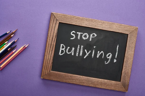 Top view of chalkboard in wooden frame with stop bullying lettering near colored pencils on purple background — Stock Photo