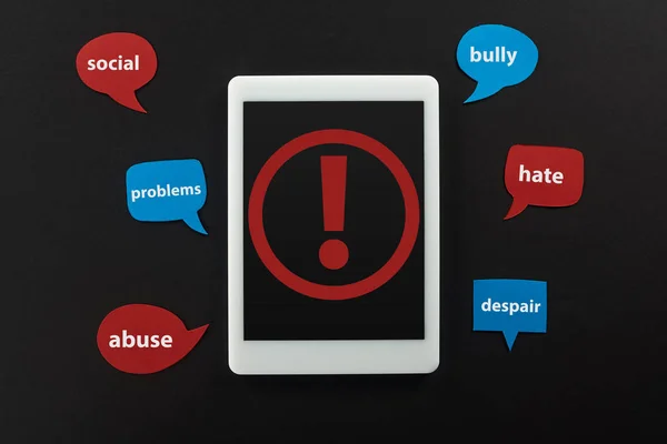 Top view of digital tablet with exclamation mark on screen on black background near speech bubbles with cyberbullying words — Stock Photo