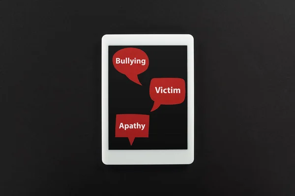 Vista superior de la tableta digital y burbujas de habla roja con bullying, palabras de víctima y apatía sobre fondo negro, concepto de cyberbullying - foto de stock