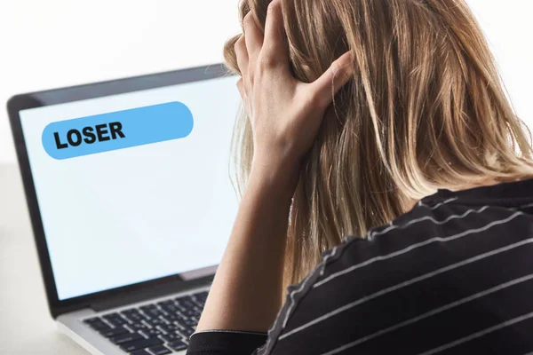 Offended blonde girl as victim of cyberbullying sitting near laptop with loser message on screen — Stock Photo