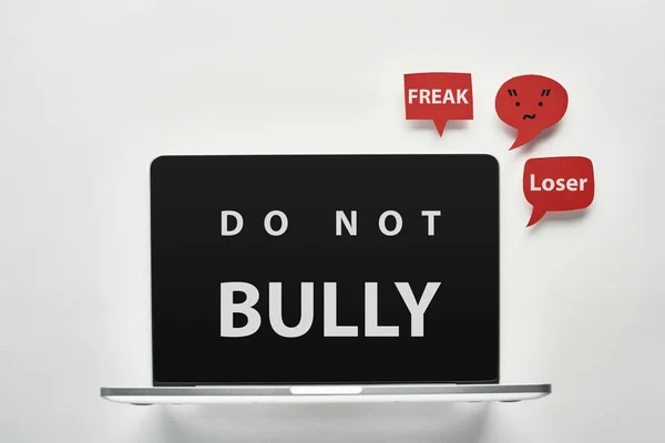 Portátil con no bully letras en la pantalla sobre fondo blanco cerca de burbujas de habla roja con palabras ofensivas, concepto de cyberbullying - foto de stock