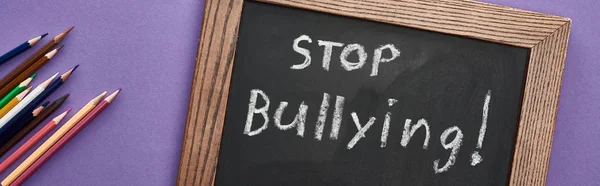 Panoramic shot of chalkboard in wooden frame with stop bullying lettering near colored pencils on purple background — Stock Photo