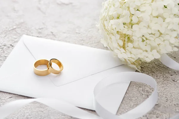 Golden rings on envelope near white ribbon and Hortense flower on grey textured surface — Stock Photo