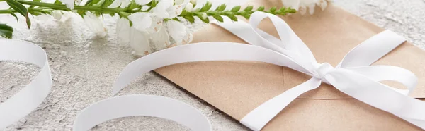 Panoramic shot of beige envelope with white ribbon near flowers on textured surface — Stock Photo