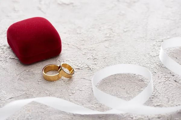 Golden rings near red gift box and white ribbon on textured surface — Stock Photo