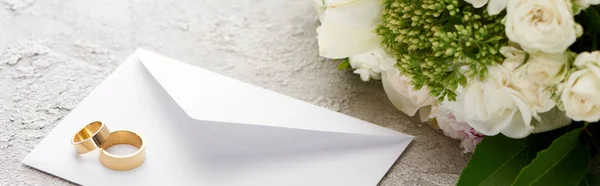 Tiro panorâmico de anéis de casamento no envelope branco perto do buquê de flores na superfície texturizada — Fotografia de Stock