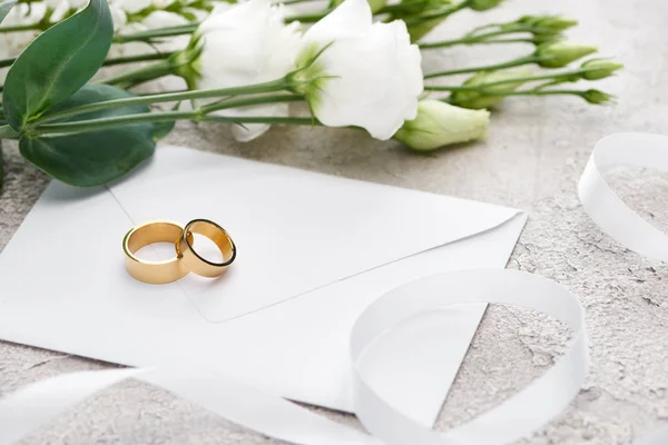 Anillos de boda dorados en sobre blanco cerca de flores de eustoma y cinta blanca - foto de stock