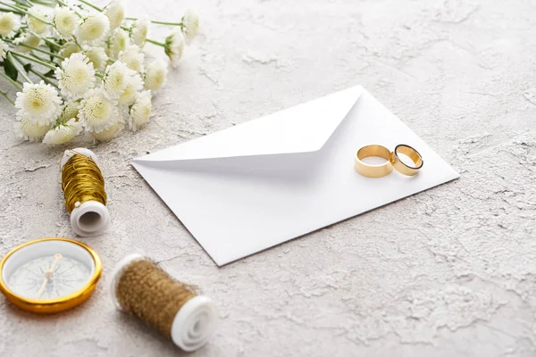Золотые кольца на белом конверте рядом с бобинсом, хризантемы и золотой компас на текстурной поверхности — Stock Photo