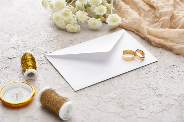 Anéis dourados no envelope branco perto de bobbins, crisântemos, pano de saco bege e bússola dourada na superfície texturizada — Fotografia de Stock