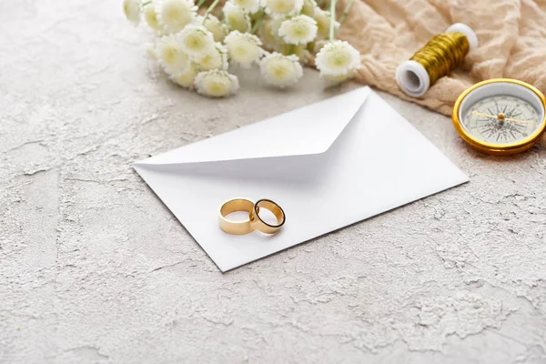 Wedding rings on white envelope near chrysanthemums, beige sackcloth, spool and golden compass on textured surface — Stock Photo