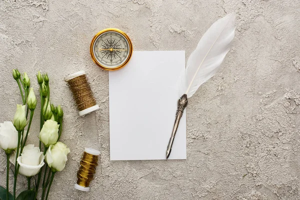 Vista superior da pena pena pena no cartão branco perto da bússola dourada, bobbins e flores brancas do eustoma na superfície texturizada cinza — Fotografia de Stock