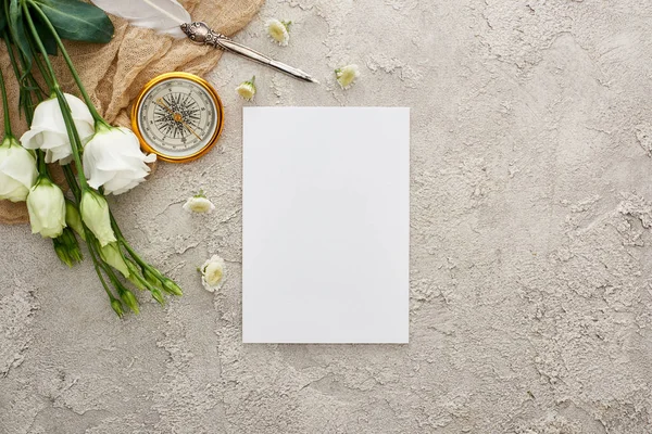 Vista superior de pluma de pluma en saco beige cerca de la tarjeta en blanco, brújula dorada y flores de eustoma blanco en la superficie texturizada gris - foto de stock