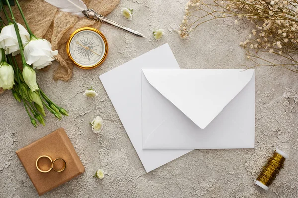 Top view of white envelope and card near beige sackcloth, compass, golden rings on gift box and white flowers on grey textured surface — Stock Photo