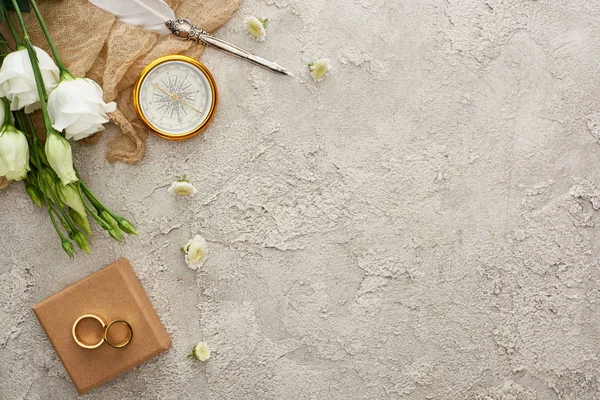 Top view of quill pen on beige sackcloth near golden compass, scattered flowers, golden rings on gift box and white eustoma flowers on grey textured surface — Stock Photo