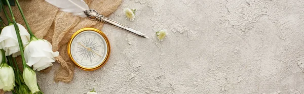 Colpo panoramico di penna d'oca su sacco beige vicino alla bussola dorata, fiori sparsi e fiori di eustoma bianco su superficie grigia testurizzata — Foto stock