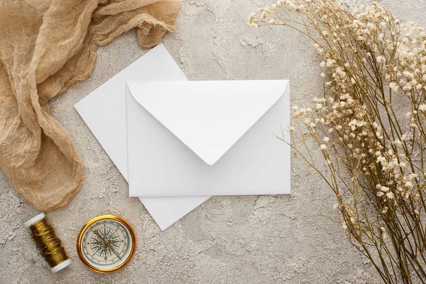 Vista superior do envelope branco e cartão perto de flores, pano de saco bege e bússola dourada na superfície texturizada — Fotografia de Stock