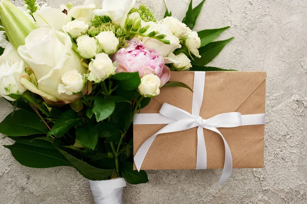 Top view of beige envelope with white ribbon near bouquet with roses on textured surface — Stock Photo