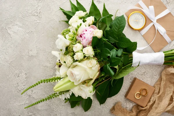 Top view of bouquet near golden compass, envelope with white ribbon, beige sackcloth and golden rings on gift box on textured surface — Stock Photo