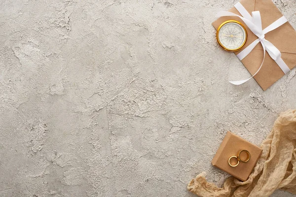 Vue du dessus de compas doré, sac beige, bagues dorées sur boîte cadeau et enveloppe avec ruban blanc sur surface texturée — Photo de stock
