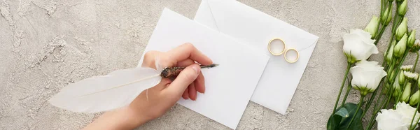 Tiro panorâmico de mulher escrevendo no cartão de convite perto do anel de casamentos e flores eustoma na superfície texturizada — Fotografia de Stock