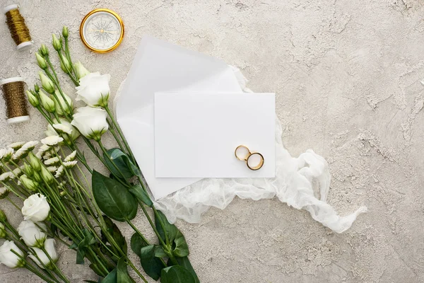 Top view of eustoma flowers near wedding rings on empty card, white cheesecloth, golden compass and bobbins on grey textured surface — Stock Photo