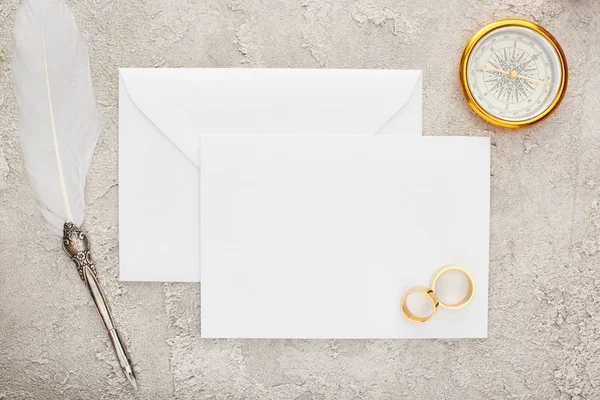 Top view of wedding rings and quill pen on white blank card and golden compass on textured surface — Stock Photo