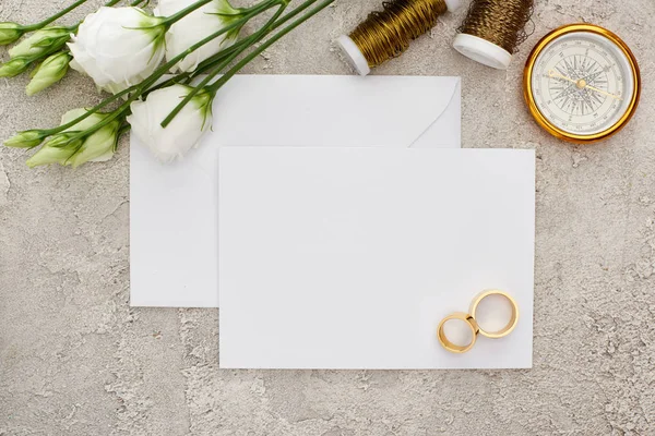 Top view of golden rings on empty card near eustoma flowers, bobbins and golden compass on grey surface — Stock Photo