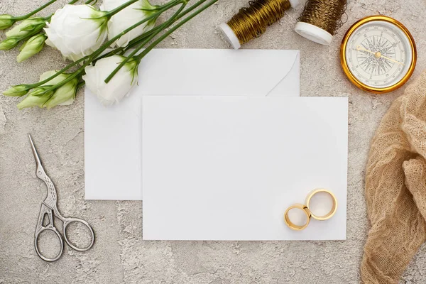 Hochzeitsringe auf leerer Karte in der Nähe weißer Eustoma-Blumen, Spulen, Scheren und goldenem Kompass auf grauer Oberfläche — Stockfoto
