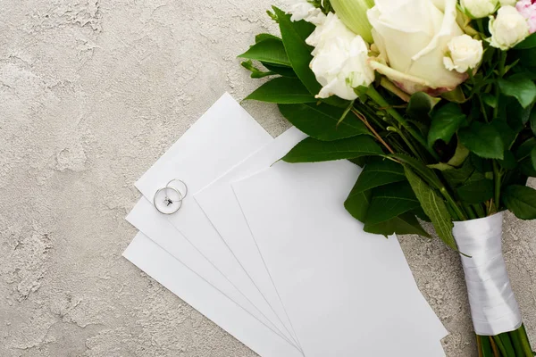 Vue du dessus des bagues argentées sur les cartes d'invitation près du bouquet sur surface texturée — Photo de stock