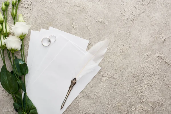 Top view of silver rings and quill pen on invitation cards near white eustoma flowers on textured surface — Stock Photo