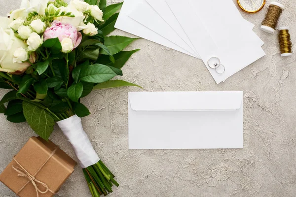 Вид зверху на білі конверти біля обручок, подарункова коробка, букет, боби і золотий компас на текстурованій поверхні — стокове фото