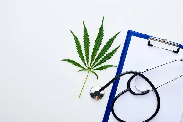 Vista dall'alto della foglia di cannabis medica vicino allo stetoscopio sugli appunti su sfondo bianco — Foto stock