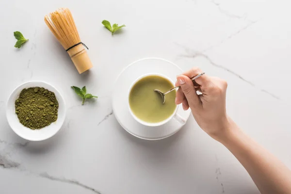 Cropped view of woman with matcha matcha tea on table with whisk, powder and mint — Stock Photo