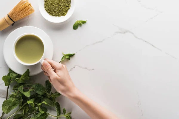 Vista dall'alto della donna che tiene il tè matcha matcha sul tavolo con frusta, polvere e menta — Foto stock