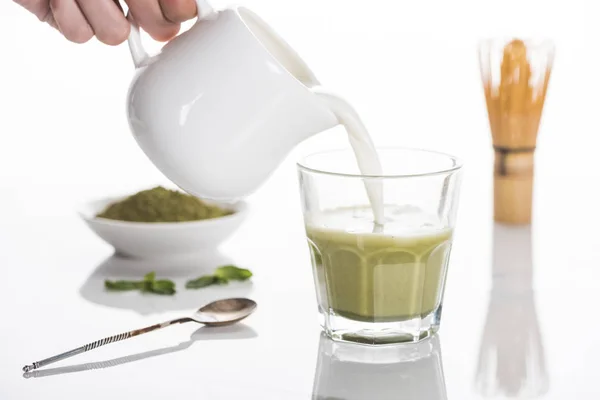 Vista recortada de la mujer vertiendo leche en el té matcha matcha en la mesa con batidor y polvo - foto de stock