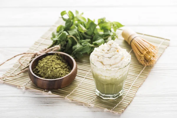 Matcha green tea with whipped cream on bamboo mat with powder, mint and whisk — Stock Photo