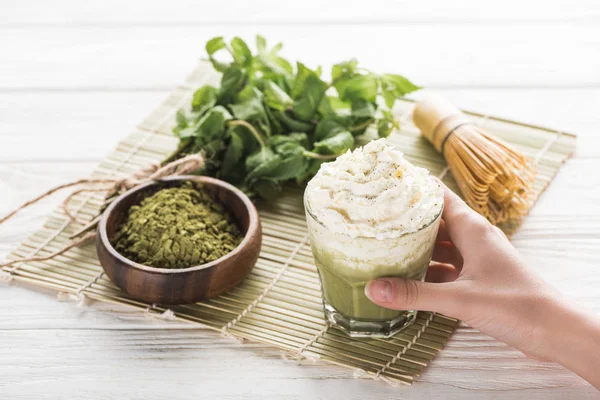 Cropped view of woman holding matcha matcha tea with whipped cream on table with whisk, powder and mint — Stock Photo