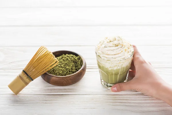 Vista recortada de la mujer sosteniendo té matcha matcha con crema batida en la mesa con batidor y polvo - foto de stock