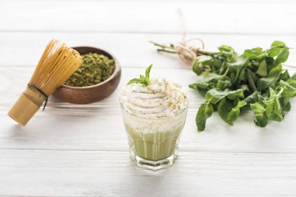 Té verde matcha con crema batida sobre mesa blanca con polvo, menta y batidor - foto de stock