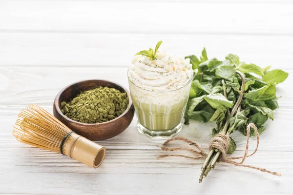 Bicchiere di tè verde matcha con panna montata su tavolo bianco con polvere, menta e frusta — Foto stock