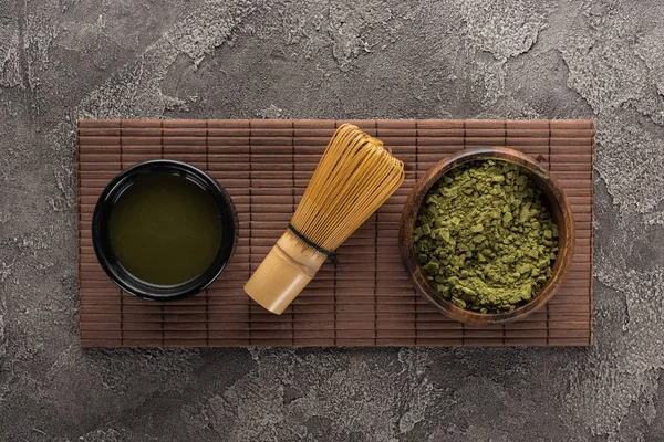 Vista superior de té matcha con batidor en estera de bambú en mesa de piedra oscura - foto de stock