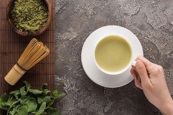 Vista ritagliata della donna che tiene il tè matcha matcha sul tavolo con frusta, polvere e menta — Foto stock