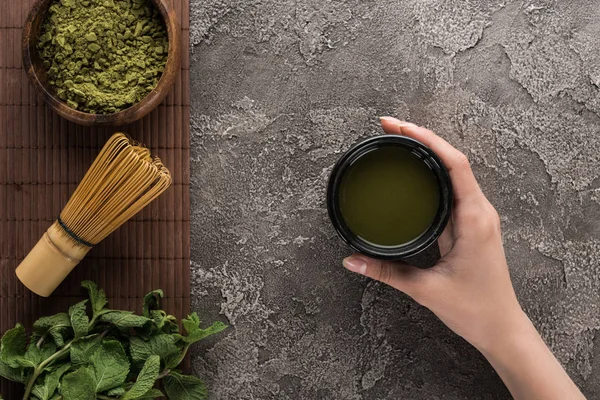 Top view of woman holding matcha matcha tea on table with whisk, powder and mint — Stock Photo