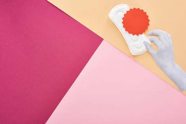 Vista dall'alto della mano bianca con asciugamano sanitario e cartellino rosso su sfondo rosa, viola e beige — Foto stock
